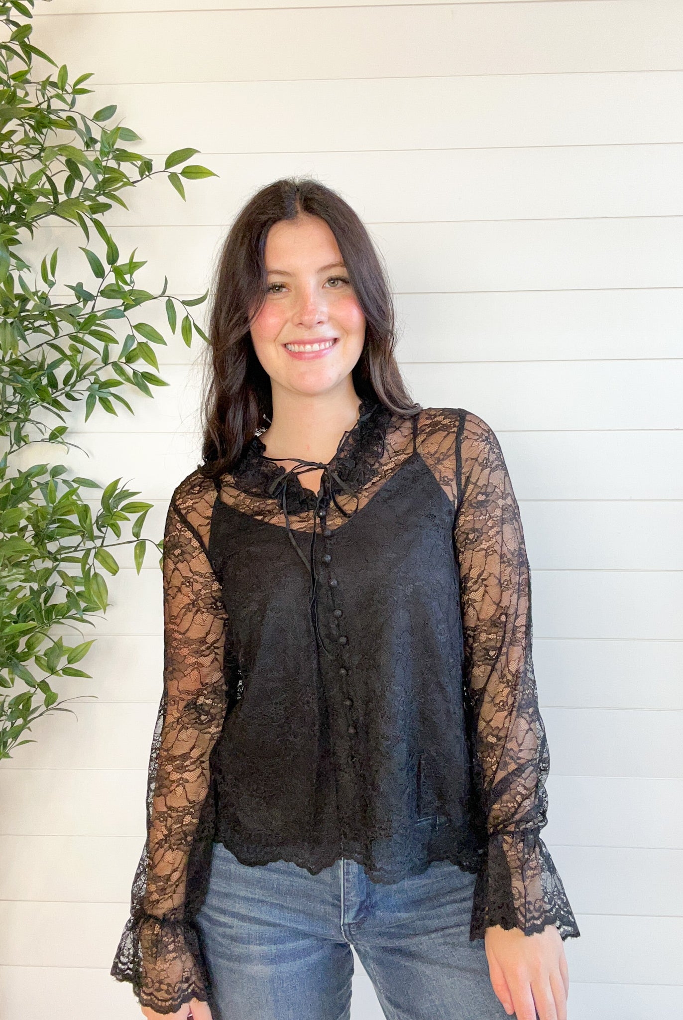 Woman wearing a black lace blouse standing next to a plant against a white wall.