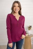 Woman wearing a purple waffle knit henley and blue jeans standing indoors.