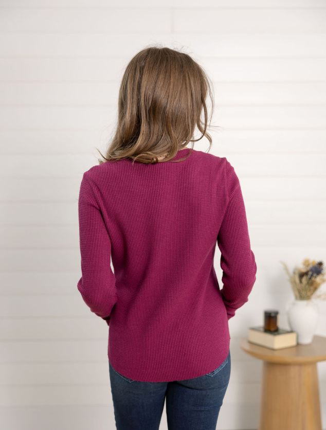 Woman wearing a purple waffle knit henley and blue jeans standing indoors.