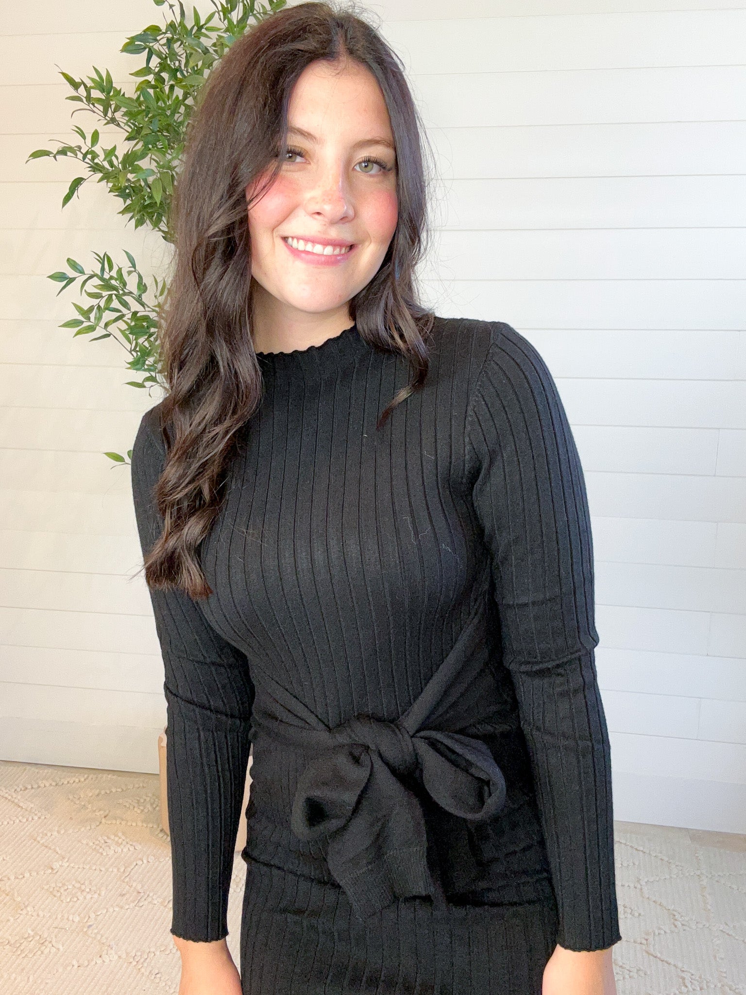 Woman wearing a black ribbed sweater with a tie detail in front of a white wall.