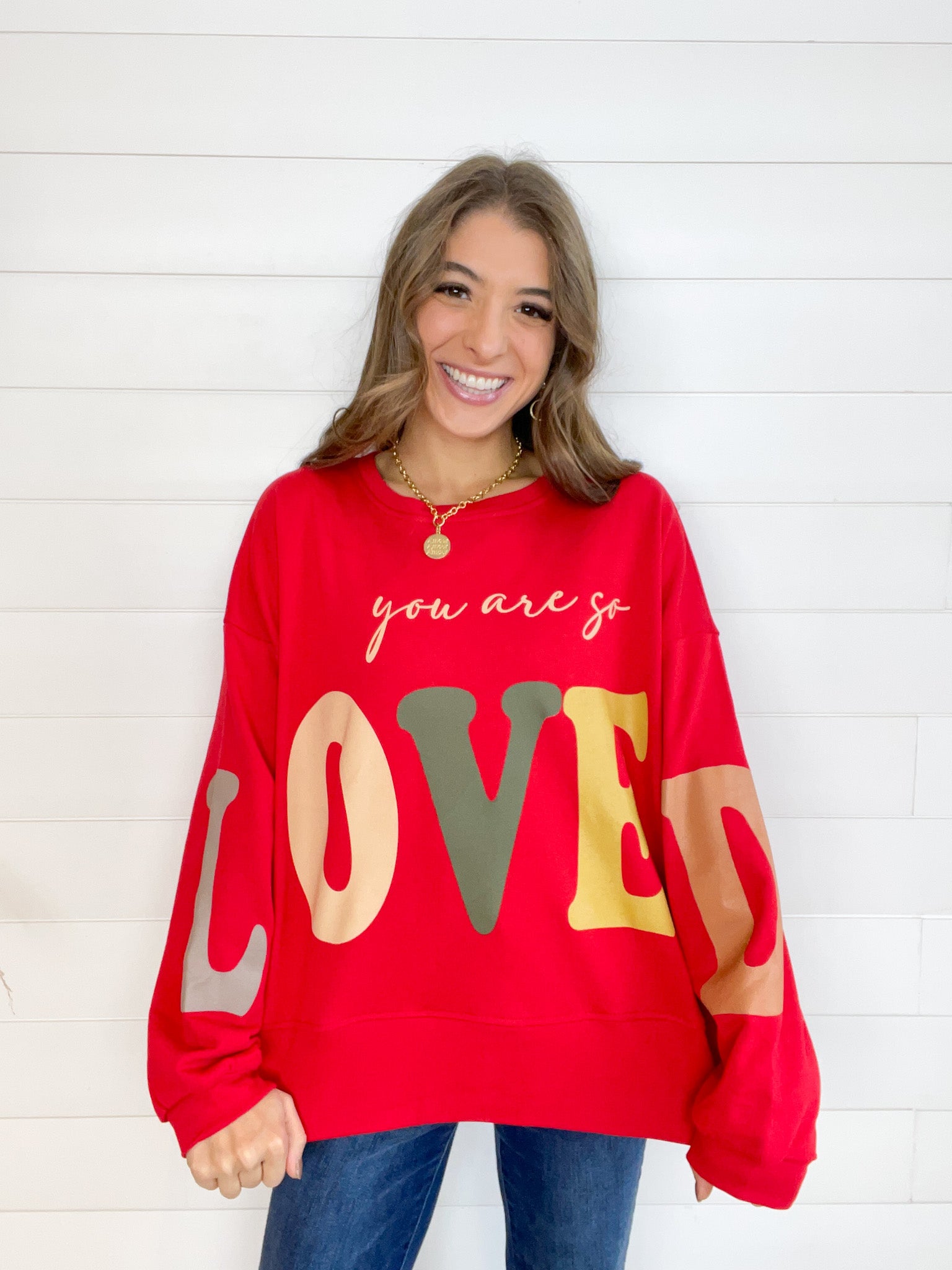 Red long sleeve sweatshirt with "You are so loved" printed across the chest, the "L" and "D" of "Loved" are on the left and right arms respectively. The word "loved" is in a warm toned multicolor print, each letter being a different color