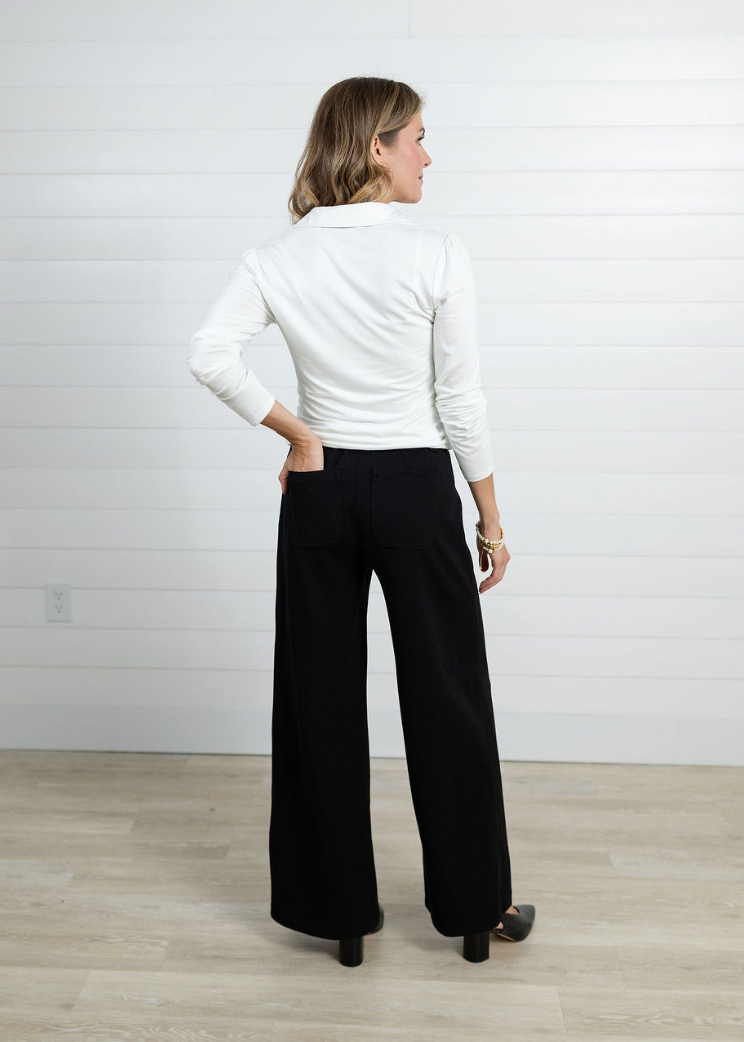 Woman wearing a white long-sleeve top and black wide-leg pants against a white wall.