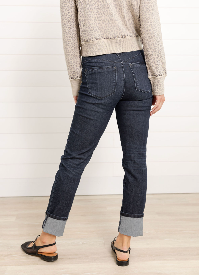 Person wearing dark blue jeans with a neutral sweater and sandals on a light wooden floor.